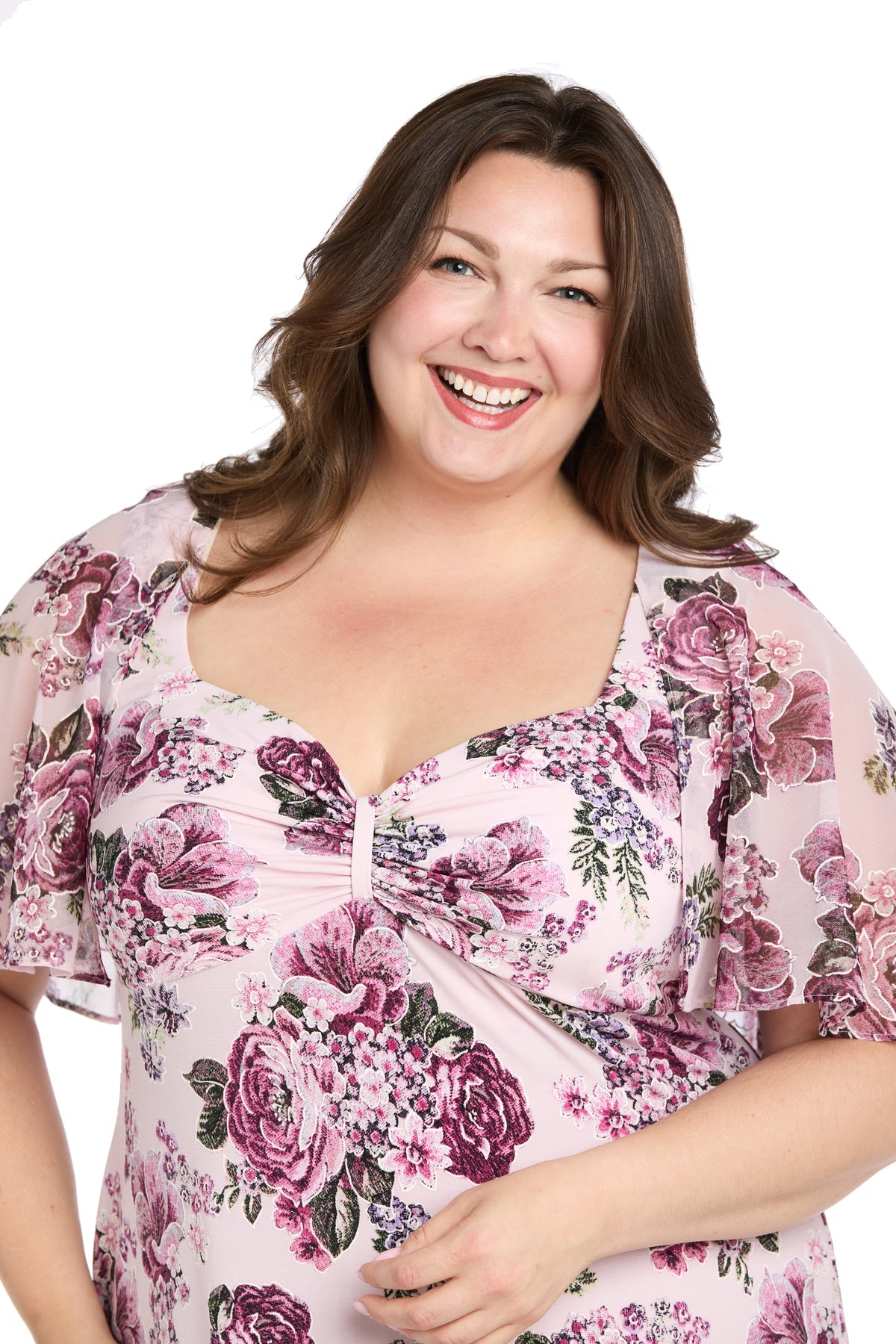 A woman with long brown hair smiles, wearing the R&M Richards 3467W plus-size pink party dress with floral pattern and sheer short sleeves, standing against a white background.